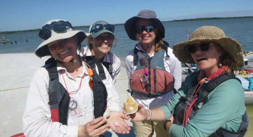 Four people wearing life jackets, sun hats and sunglasses stand on a beach. Two of them are holding small seashells. 